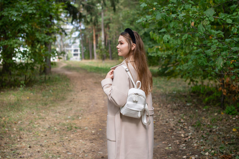 A student girl walks with a backpack and in a long coat on the road in the forest