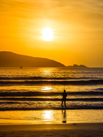 Silhouette person fishing while standing in sea against sky during sunset