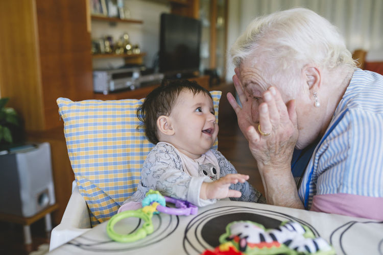 Happy great-grandmother playing with a baby girl sitting in the high chair next to the table with toys