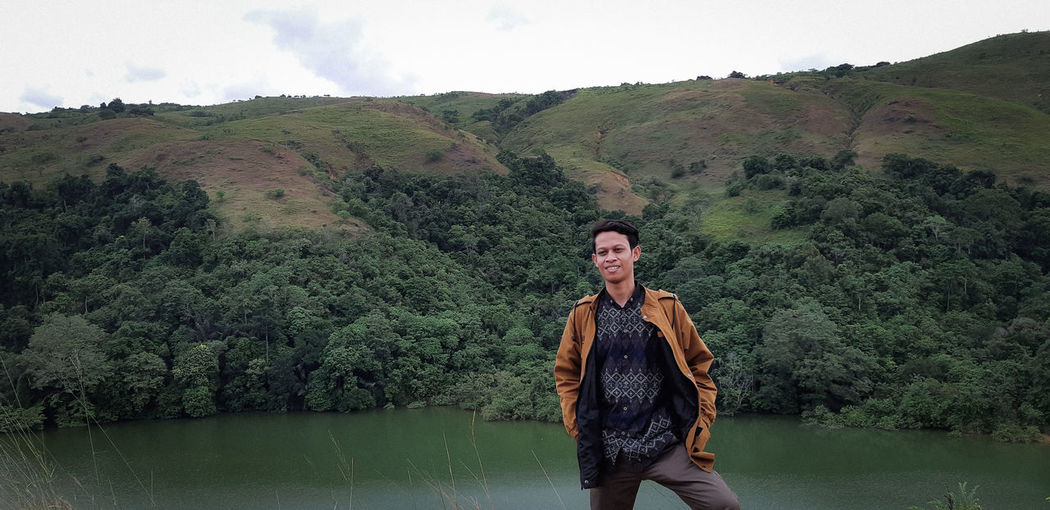 Smiling young man standing against river and mountain in forest