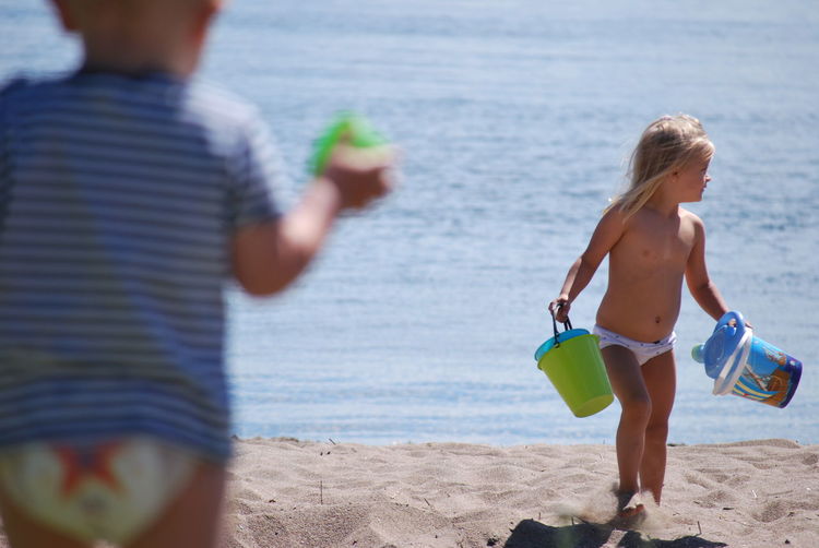 Shirtless girl holding buckets while walking on shore at beach during sunny day