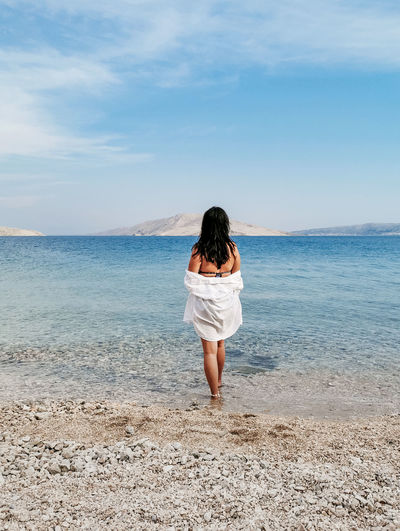 Rear view lifestyle image of stylish young woman in white shirt standing on beach and looking at sea