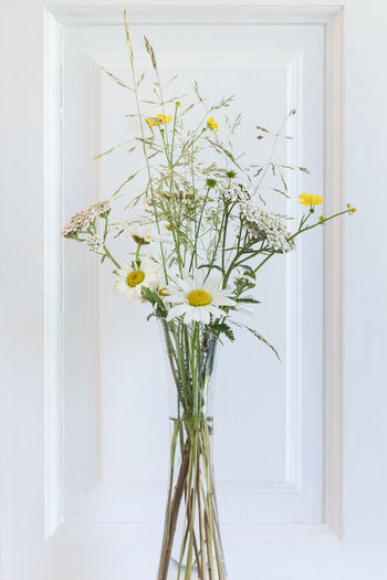 Bunch of wild summer flowers and grass in transparent vase in front of white cabinet door