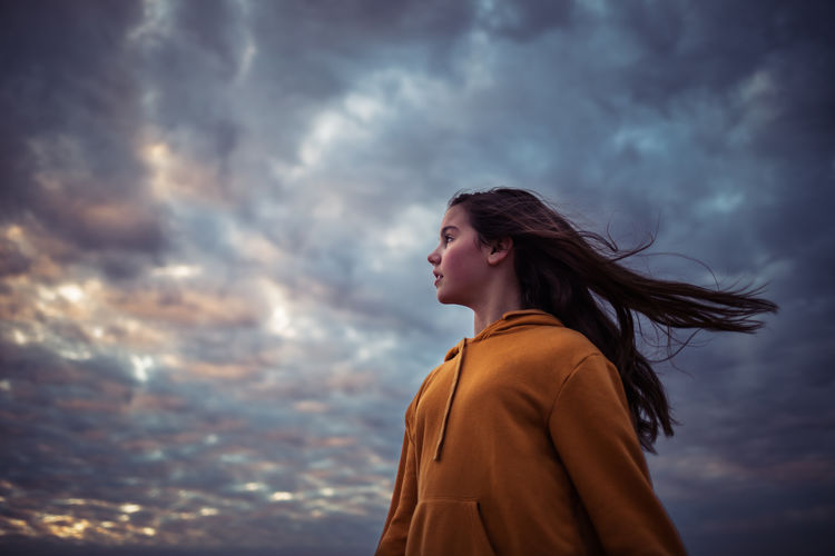 Low angle view of girl with long hair looking away while standing against cloudy sky