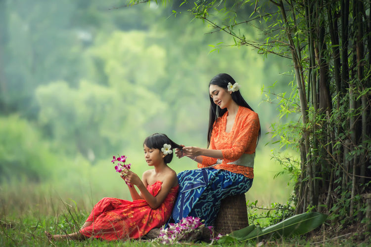 Mother braiding daughter hair while sitting on field against trees
