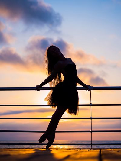 Silhouette woman dancing by railing against sea during sunset