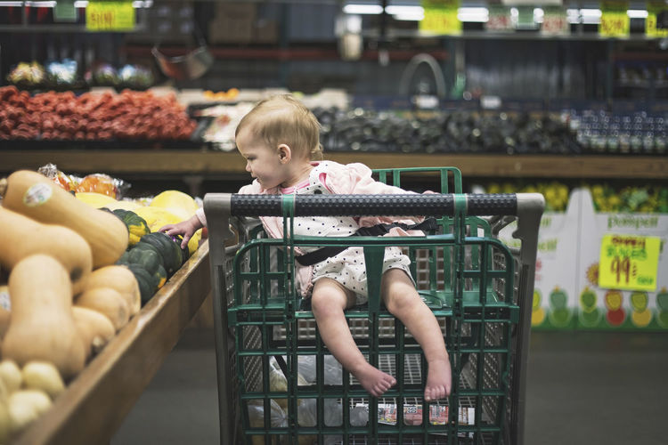 Full length of baby girl sitting on shopping cart while picking vegetables from market stall at supermarket