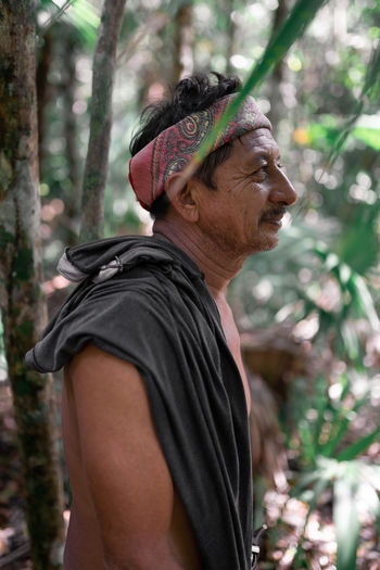 Side view of shirtless ethnic male worker in colorful headband standing in forest with tall trees during work in nature