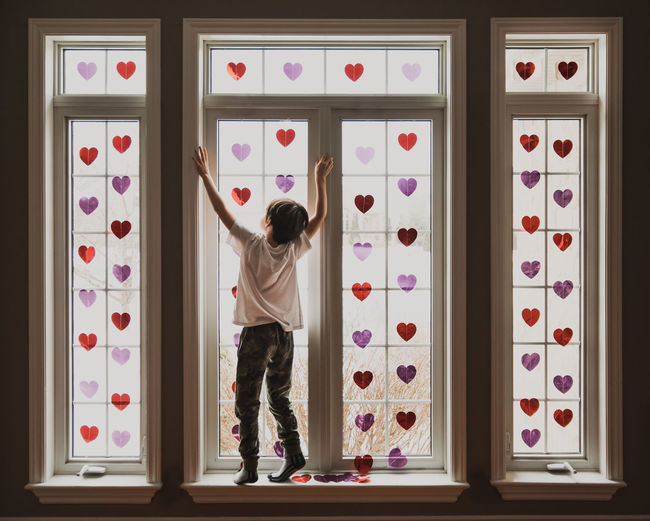 Boy standing on windowsill of windows covered in small shiny hearts.