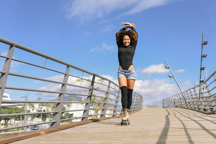 Portrait of woman with arms raised wearing roller skates on footbridge in city against sky