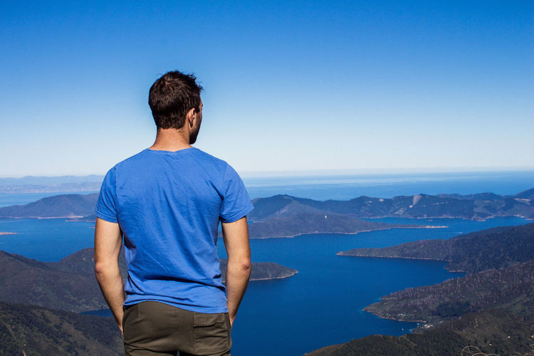 Rear view of man looking at sea while standing on mountain against blue sky