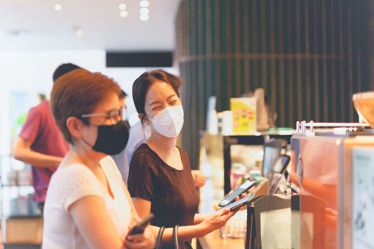 Two happy female friends in protective mask buying coffee in cafe.