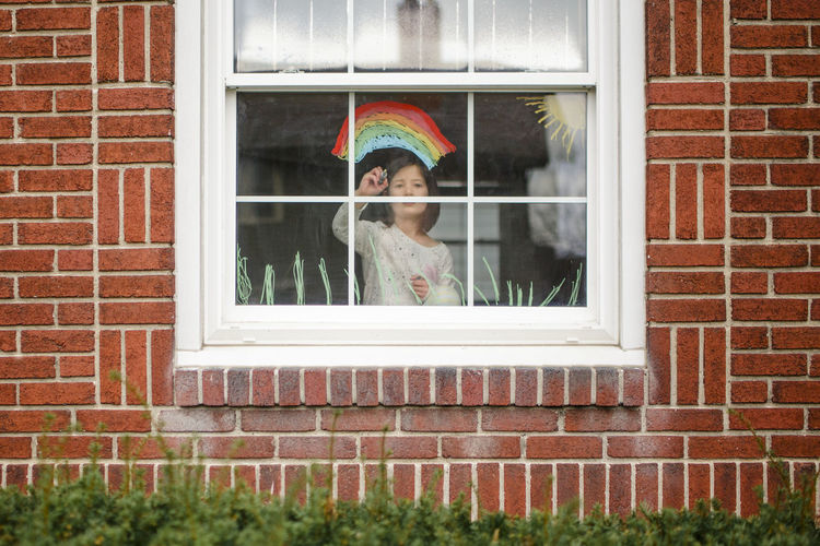View from outside of small child standing in window drawing a rainbow