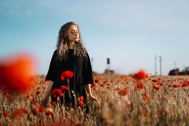 Beautiful young woman standing by poppy flowers on field against sky