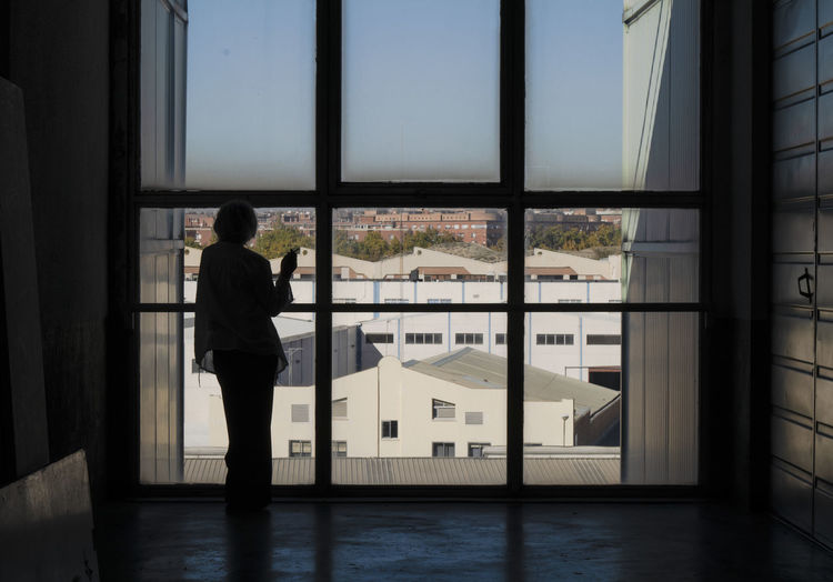 Silhouette of mature woman smoking at glass window looking at view of industrial zone