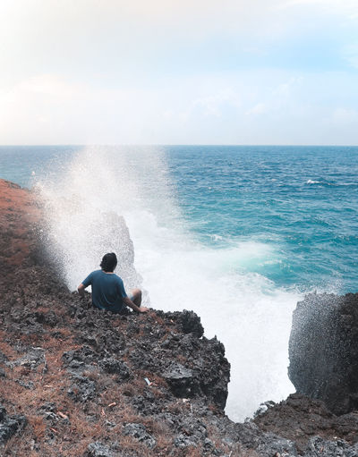 Rear view of man looking at sea while sitting on rock