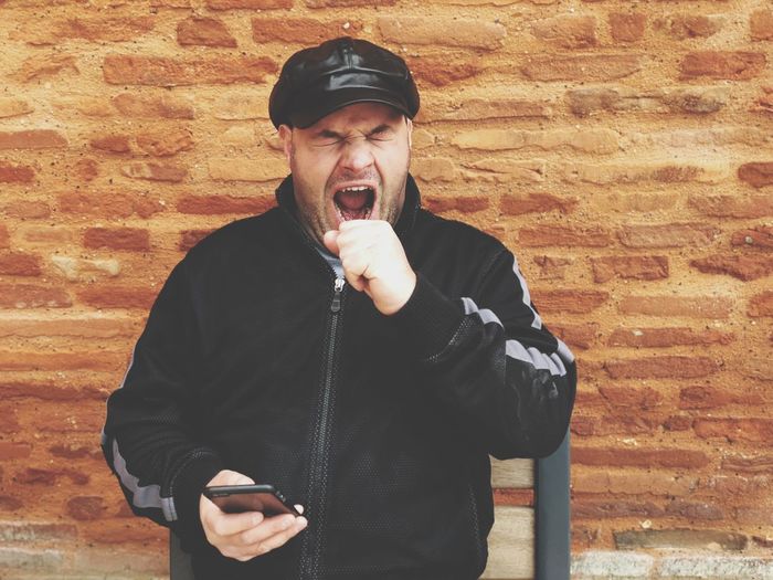 Man holding smart phone while yawning against brick wall