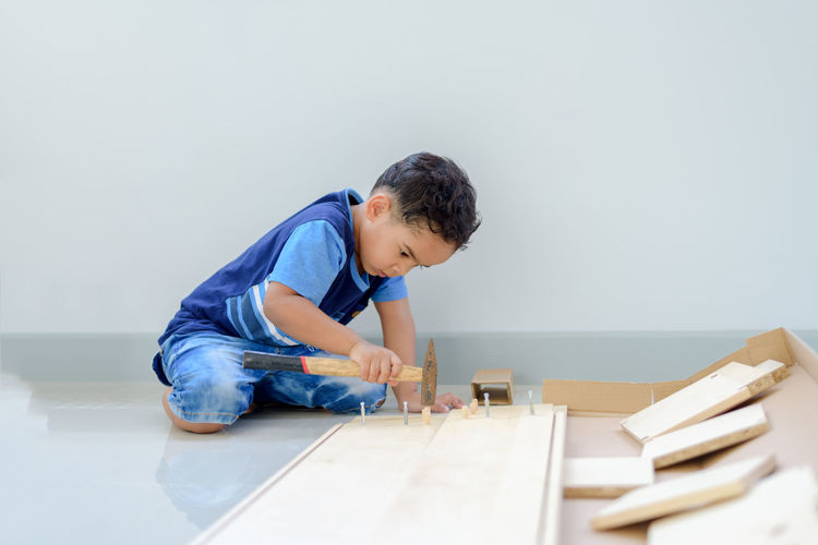 Boy hammering nails on wooden plank while sitting on floor at home