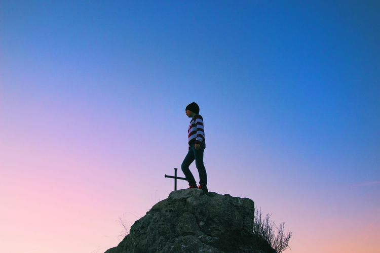 Low angle view of boy standing on rock against clear sky during sunset