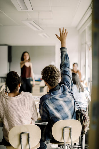 Male student with hand raised while female teacher pointing in classroom
