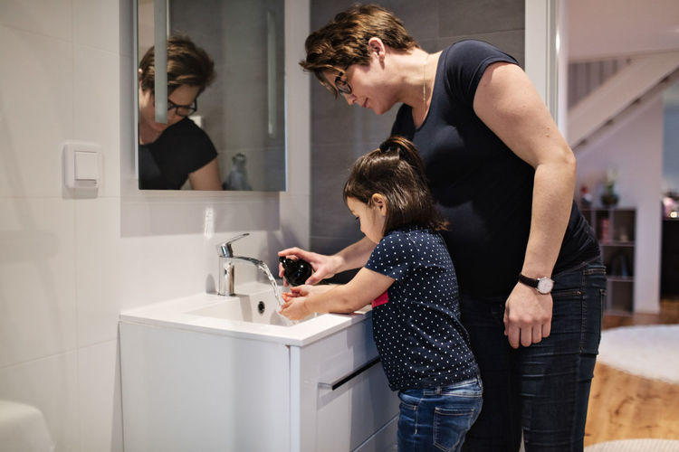 Woman assisting daughter in washing hands at sink in house