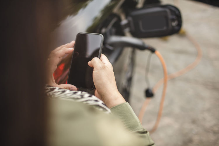 Midsection of woman using smart phone while standing by charging electric car