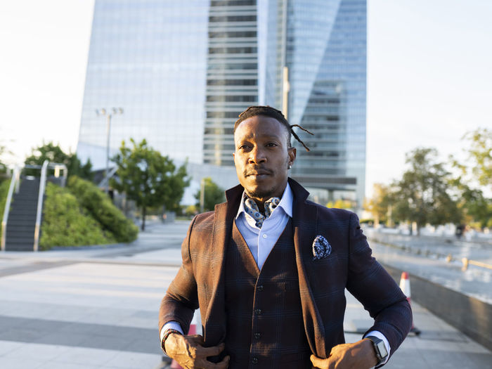 Serious african american male entrepreneur wearing classy suit standing with hand in pocket on street in city center and looking away