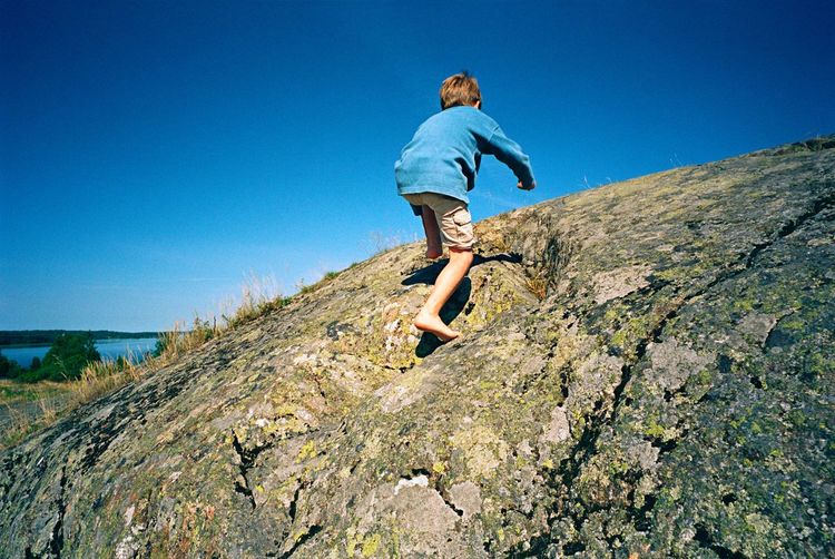 Low angle view of boy on rock against clear blue sky