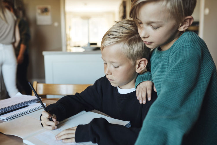 Boy with hand on shoulder of brother doing homework in book at home
