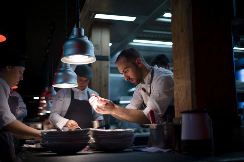 Young cooks men in aprons carefully serving meal in white ceramic dishes on restaurant kitchen