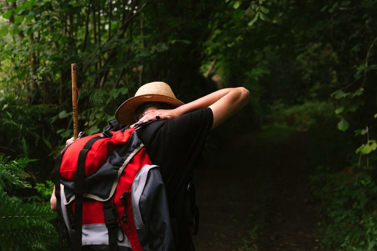 Back view of female tourist with backpack standing in dark woods in summer and resting during vacation