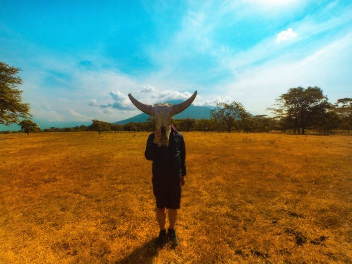 Woman covering face with animal skull while standing on land against sky