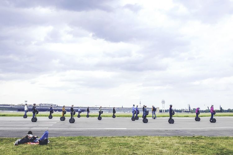 People riding segway on road against cloudy sky with women lying on grass