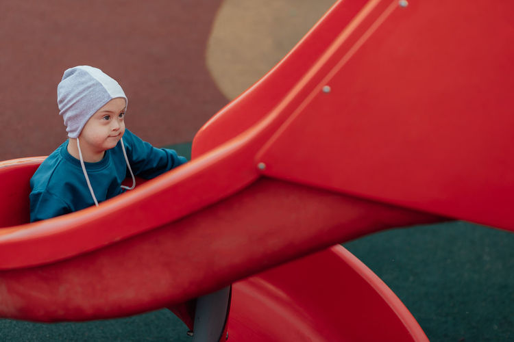Cute little boy with down syndrome in a funny hat walks in the playground, children's slide