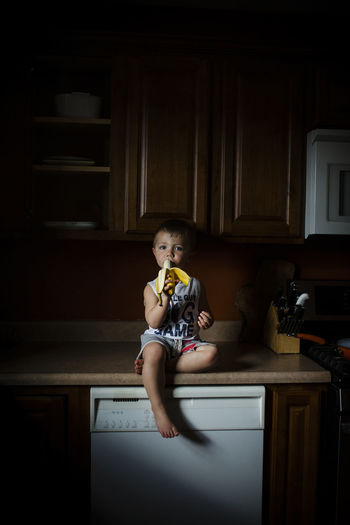 Portrait of boy eating banana while sitting on kitchen counter at home
