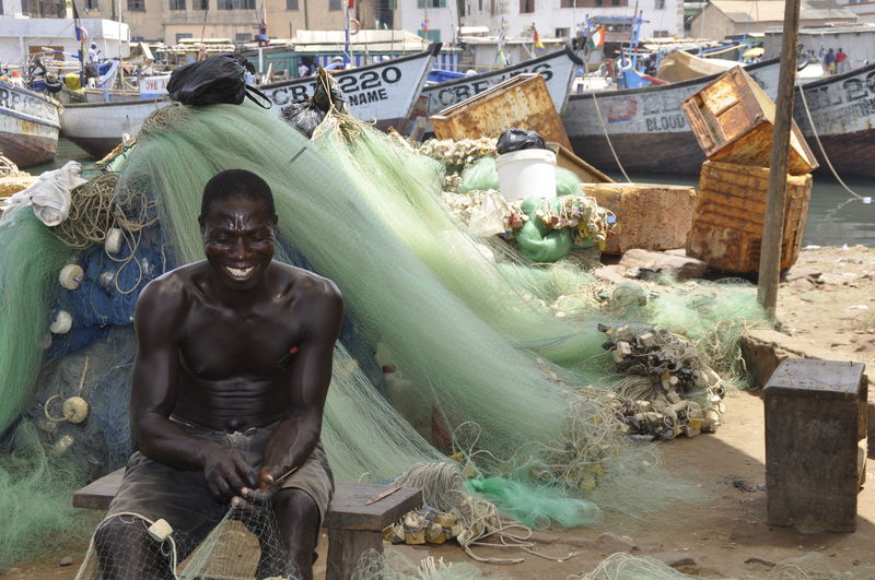 Smiling shirtless man holding fishing net while sitting on bench