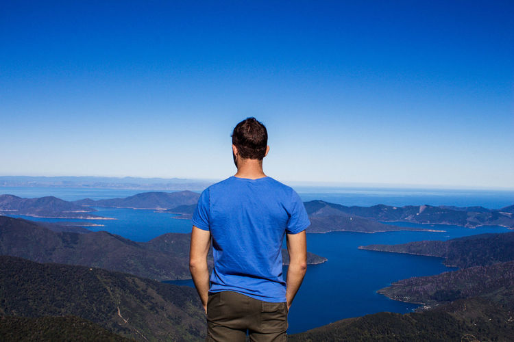 Rear view of man looking at mountains and sea against clear blue sky