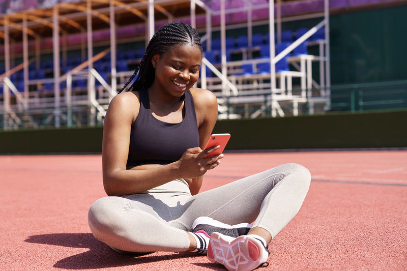 Cheerful black woman in sportswear sits on red track on stadium checking social networks on phone