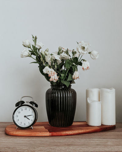 Flowers in vase by clock and candles on table against white wall
