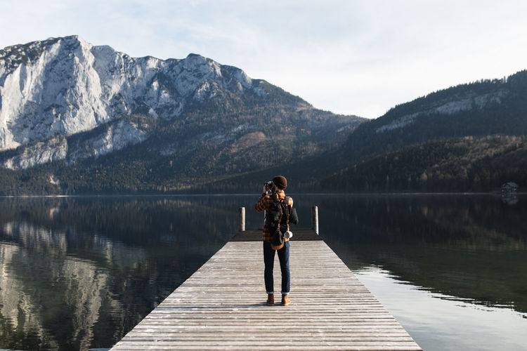 Back view of unrecognizable male tourist in warm outfit standing on wooden boardwalk and taking picture of amazing scenery of lake surrounded by rocky mountains in autumn day