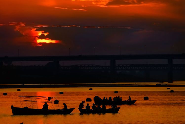 Silhouette people sitting on rowboats in river against sky during sunset