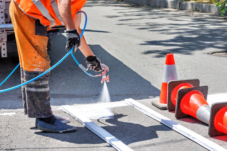 A road worker paints a white road marking of a pedestrian crossing with an airbrush on a sunny day.