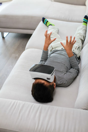High angle view of boy wearing virtual reality simulator while lying on sofa at home