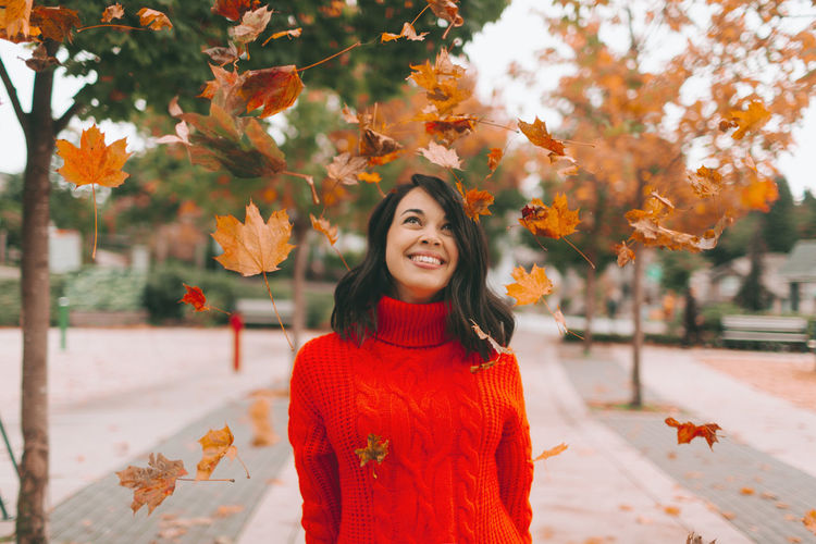Smiling woman looking at leaves while standing on footpath during autumn