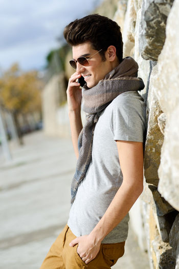 Side view of young man talking on smart phone while standing by wall