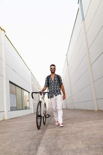 Low angle of delighted male in trendy wear walking with modern bike along narrow street and looking forward
