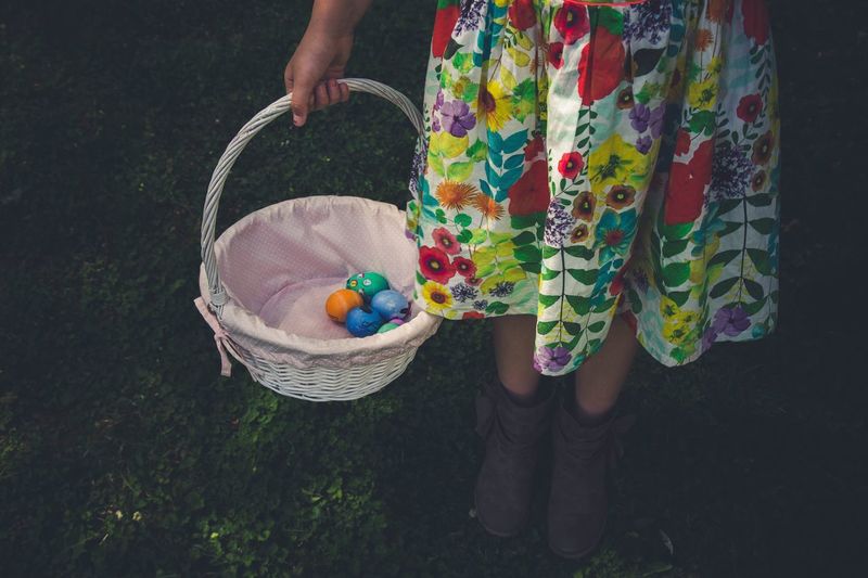 Low section of girl holding easter eggs in wicker basket while standing on grassy field