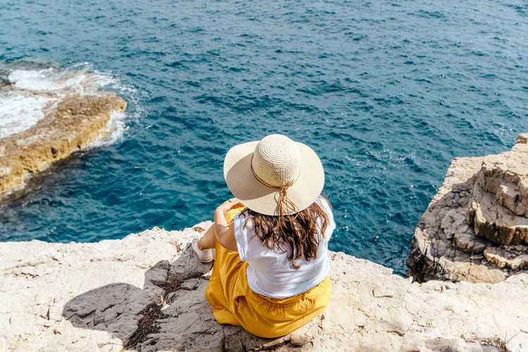 Young woman in summer clothes and hat sitting on edge of cliff above sea.