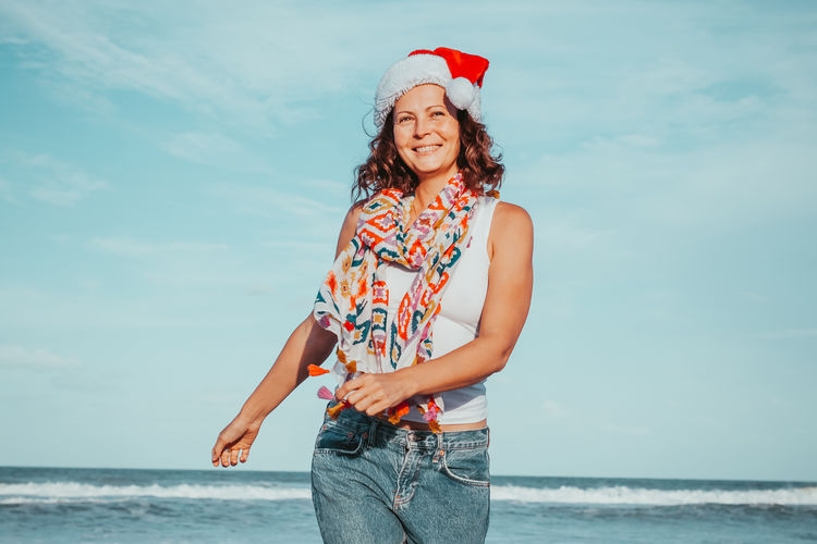 Happy suntan young woman in santas hat and colorful casual clothing walking on the beach. christmas 