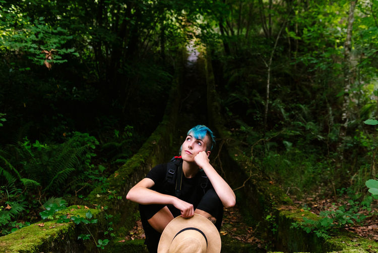 Relaxed traveling female with blue hair sitting in mossy woods during vacation and dreaming while looking up